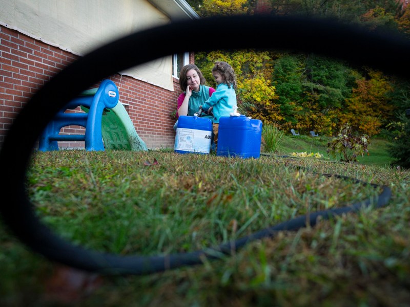 A woman and a child sit outside near a brick house, with blue containers beside them; a black hose curves in the foreground, and trees with autumn foliage are in the background.