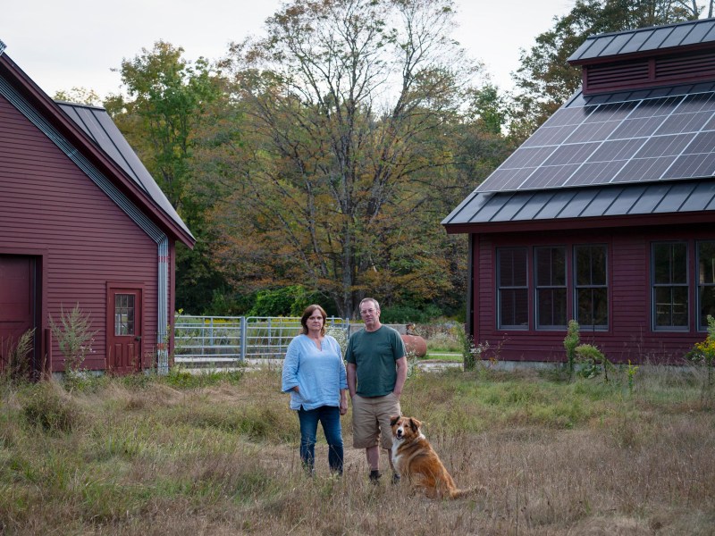 A woman and a man stand with a dog on grass between two red barns, one of which has solar panels on the roof. Trees with fall foliage are visible in the background.
