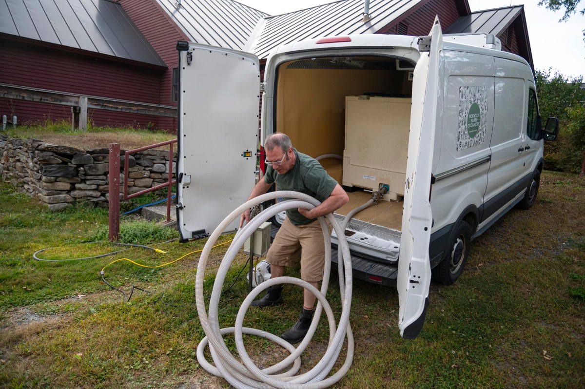A man unloads a large hose from the back of a white utility van parked on grass near a red barn and stone wall.