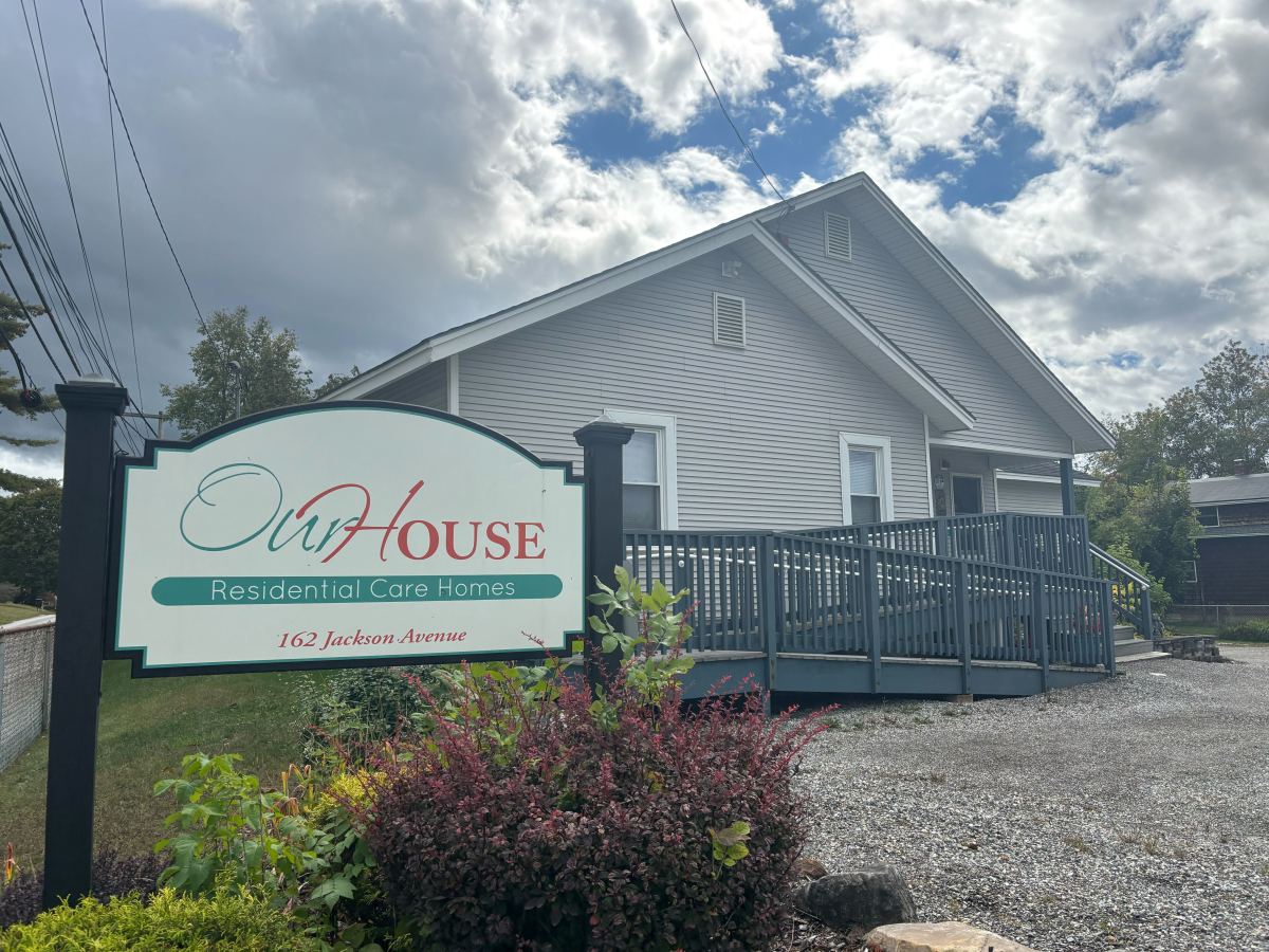 A single-story gray house with a ramped deck is shown behind a sign reading “Our House Residential Care Homes, 162 Jackson Avenue.” Shrubs and gravel are in the foreground.