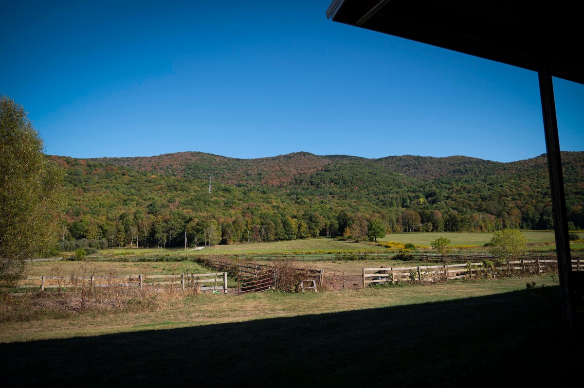 A rural landscape with green hills, fields, and a wooden fence under a clear blue sky; part of a roof casts a shadow in the foreground.