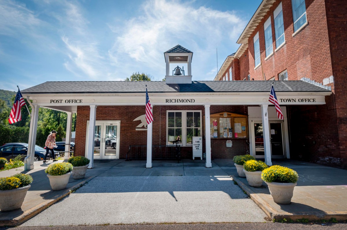 A brick building with signs for the Richmond Post Office and Town Office, two U.S. flags, potted plants, and a bell tower above the entrance.