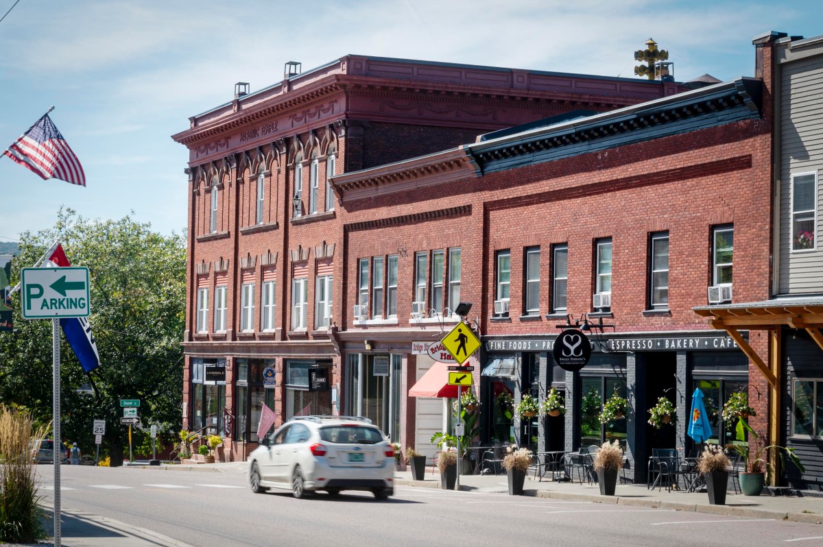 A white car drives past red brick buildings with shops and outdoor seating on a sunny day in a small town. An American flag and a parking sign are visible.