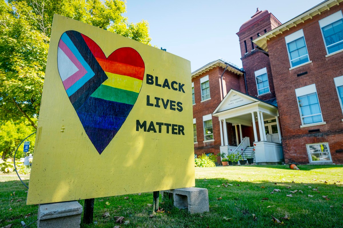 A yellow sign with a rainbow heart and the words "Black Lives Matter" stands in front of a red brick building on a sunny day.