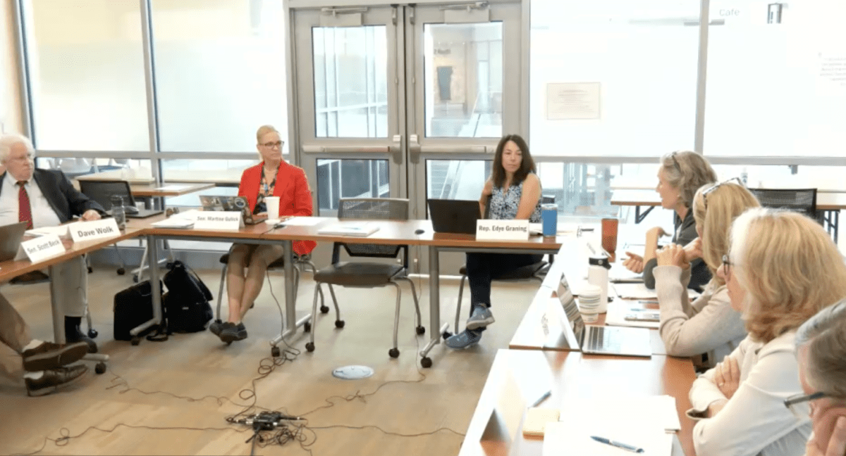 Six people sit around tables in a conference room, engaged in discussion, with laptops, papers, and coffee cups in front of them.