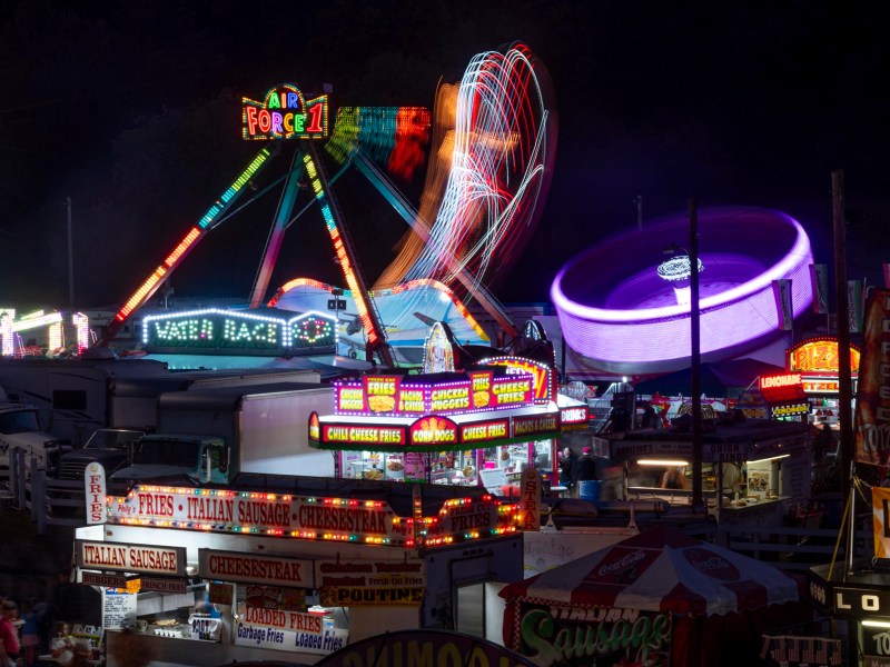 Nighttime scene at a fair with brightly lit food stalls and colorful amusement rides in motion, including a spinning ride and a Ferris wheel.