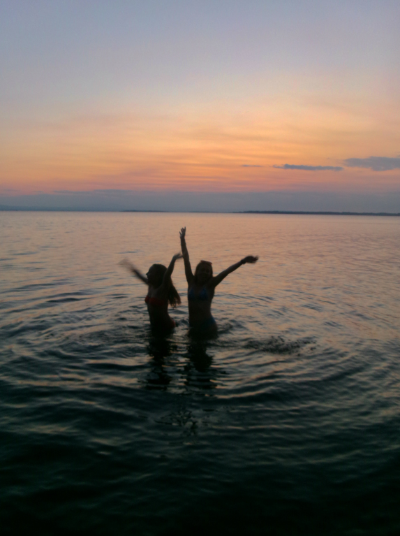 Two people stand in calm water at sunset, raising their arms as gentle waves ripple around them; the sky is orange and purple.