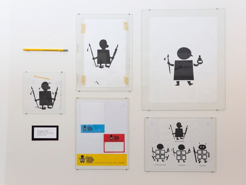 Various framed prints of simple black robot-like figures are displayed on a white wall, along with a pencil and a colorful print at the bottom center.
