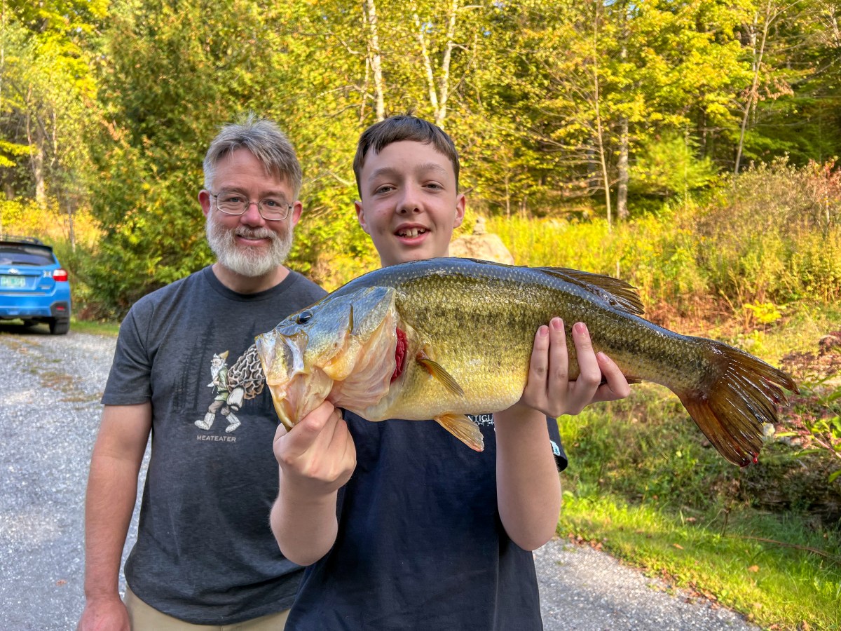 A boy holds a large fish while standing next to an older man on a gravel path, with trees and a blue car in the background.