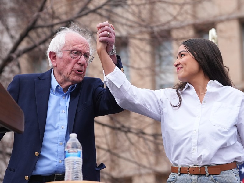 An older man in a suit and a younger woman in a white shirt hold hands raised together on an outdoor stage, with trees and a building in the background.