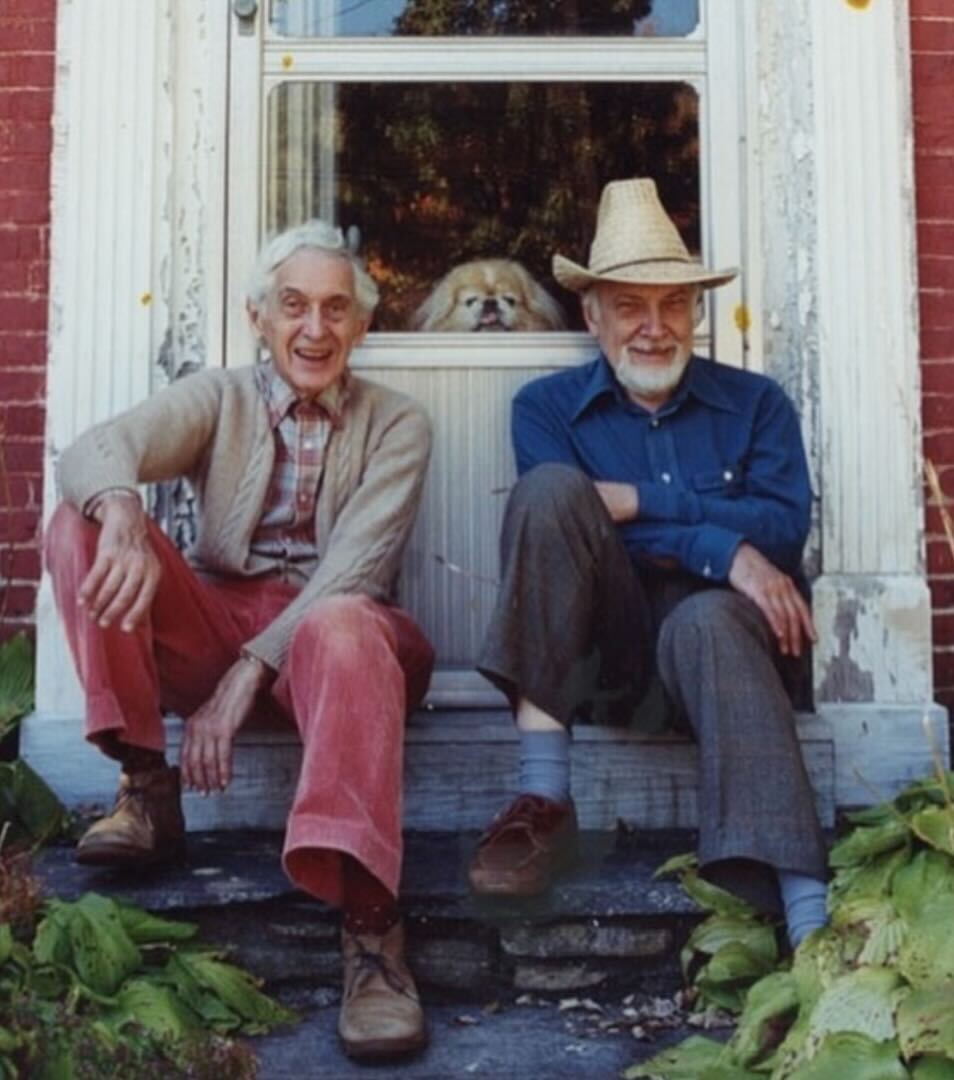 Two elderly men sit on porch steps in front of a doorway, smiling. A dog looks out from behind the glass of the closed door behind them.