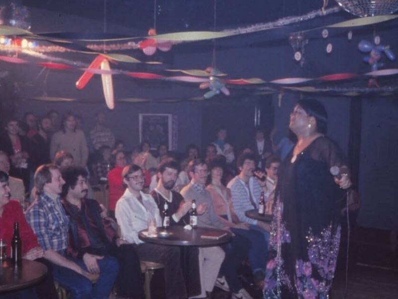 A performer sings on stage in front of a seated audience at a dimly lit nightclub decorated with streamers and balloons.