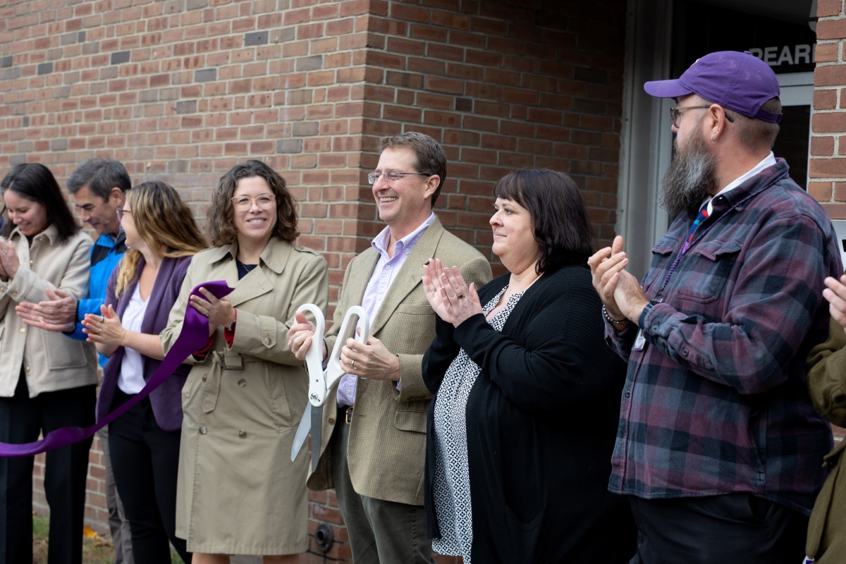 A group of people stand in front of a brick building at a ribbon-cutting ceremony. One person holds large scissors, while others clap and smile.