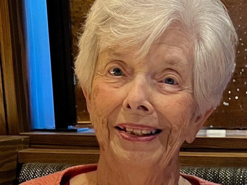 A woman with short white hair, wearing a coral pink top, smiles while seated indoors at a restaurant.