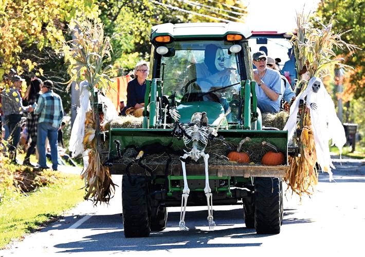 A green tractor decorated with skeletons, pumpkins, and corn stalks drives down a street during a fall or Halloween parade, with several people riding on it.