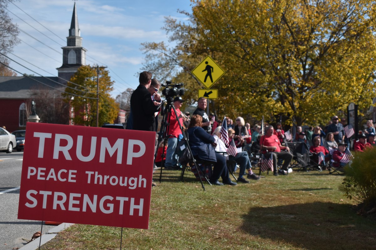 A group of people sit and stand outdoors near a red sign reading "TRUMP PEACE Through STRENGTH" with a church and fall trees in the background.
