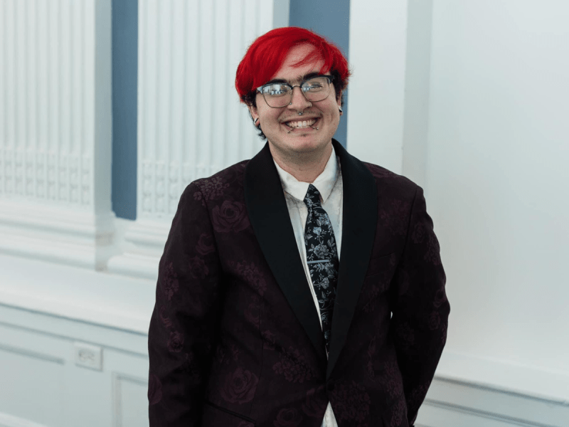 A person with red hair, glasses, and a patterned suit jacket stands smiling in front of a white and blue ornate wall.
