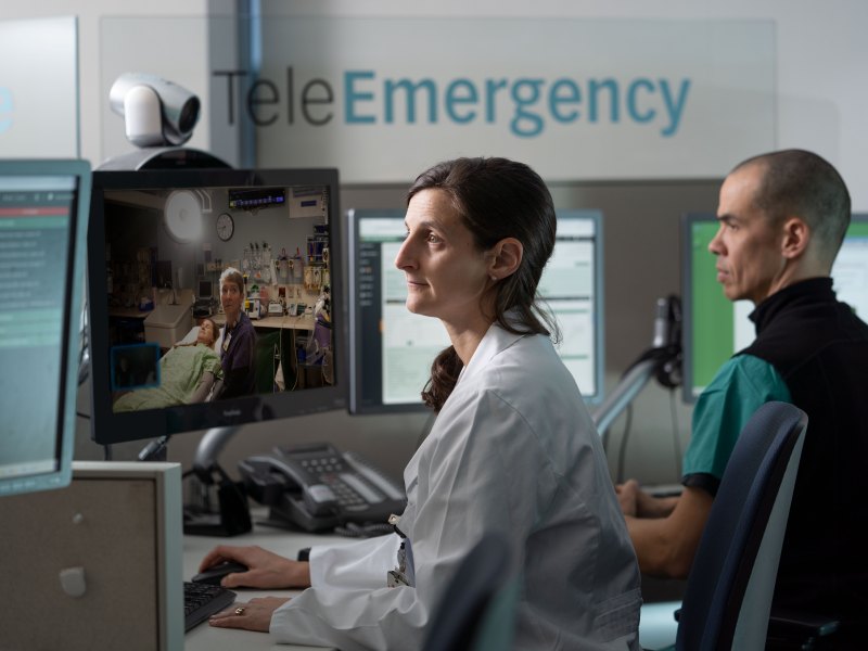 Two medical professionals work at computer stations, conducting a telehealth video call with a patient in a hospital room. "TeleEmergency" is visible on the wall behind them.