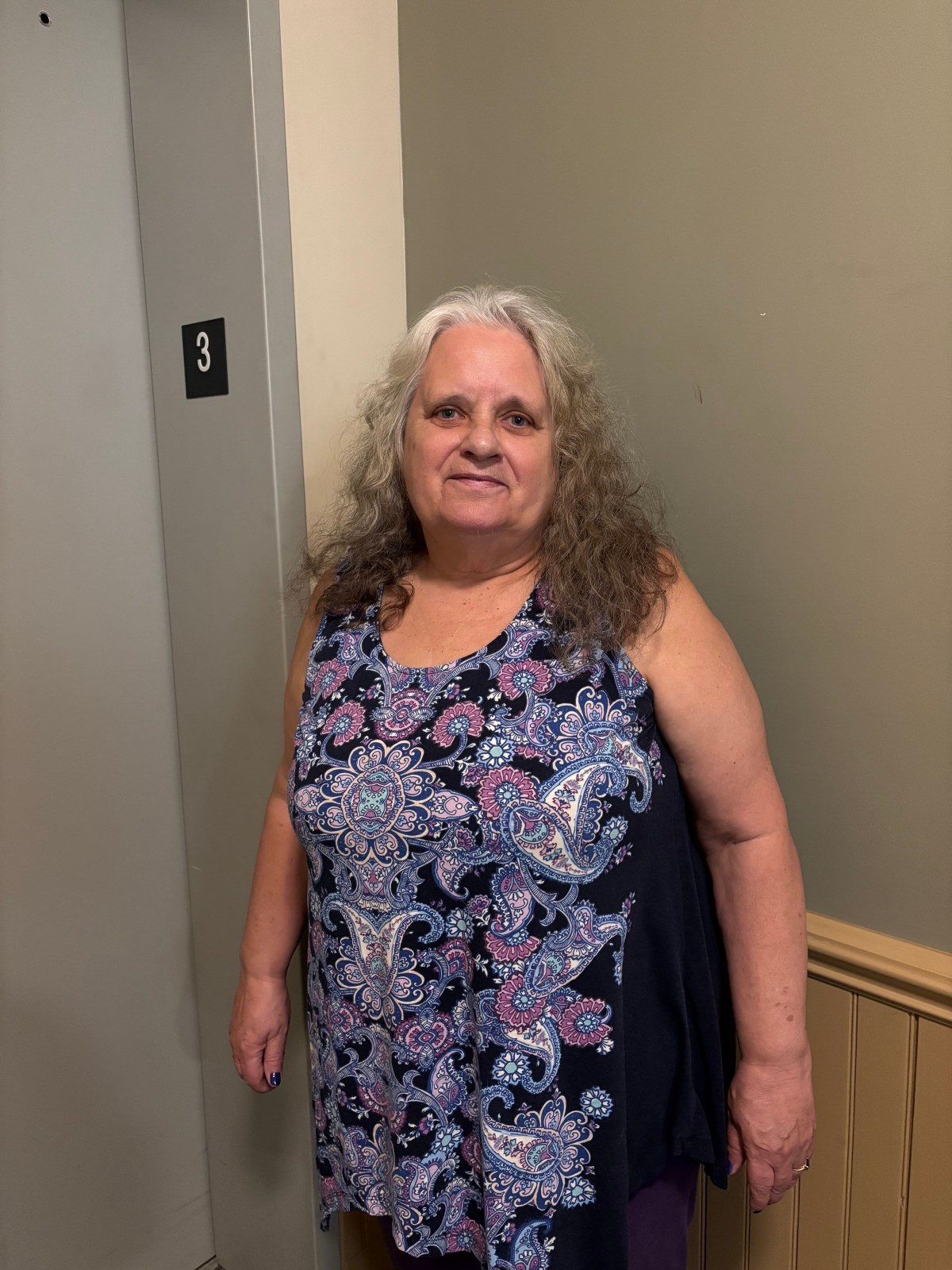 A woman with long gray hair wearing a patterned sleeveless top stands indoors next to an elevator marked with the number 3.