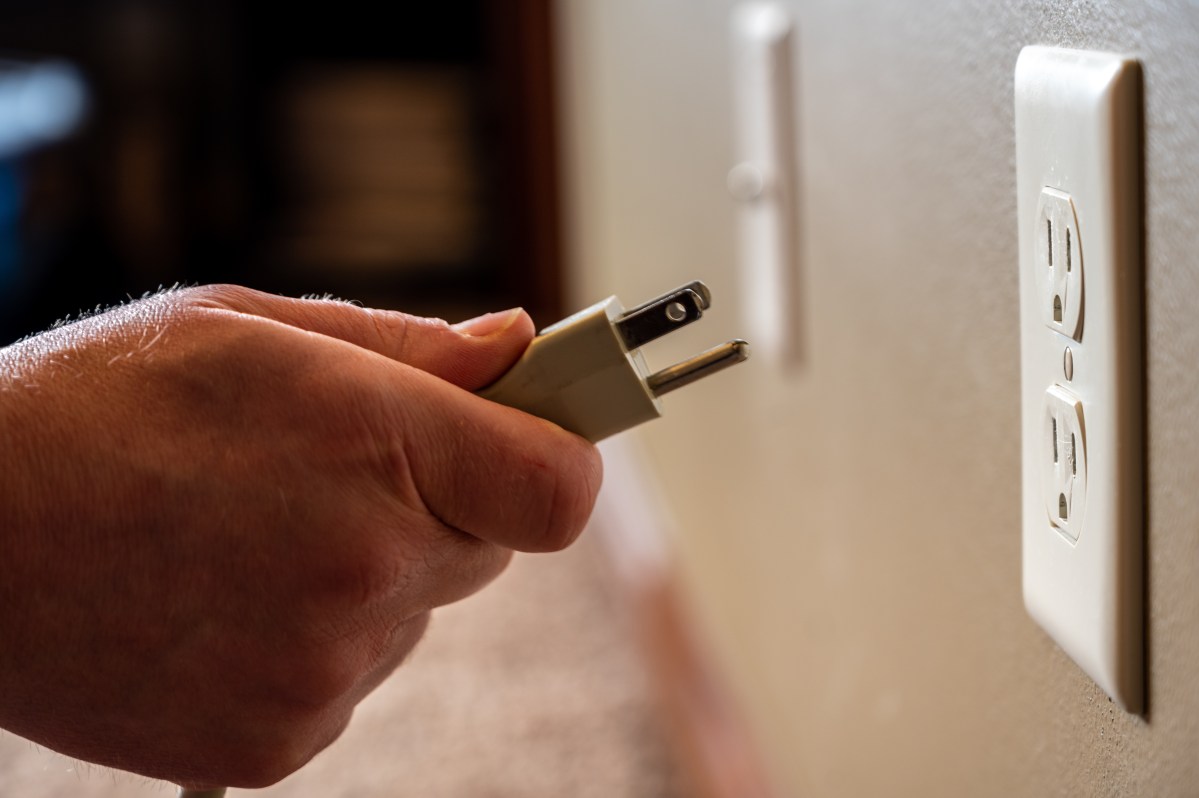 A hand holds an electrical plug close to a wall outlet, preparing to plug it in.