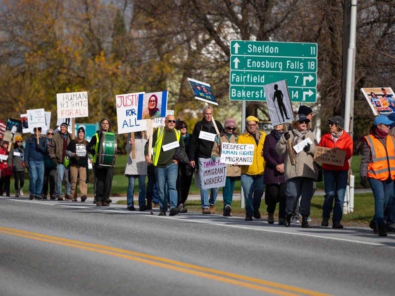 A group of people march on a roadside holding signs advocating for human rights and immigrants, with road signs and trees in the background.