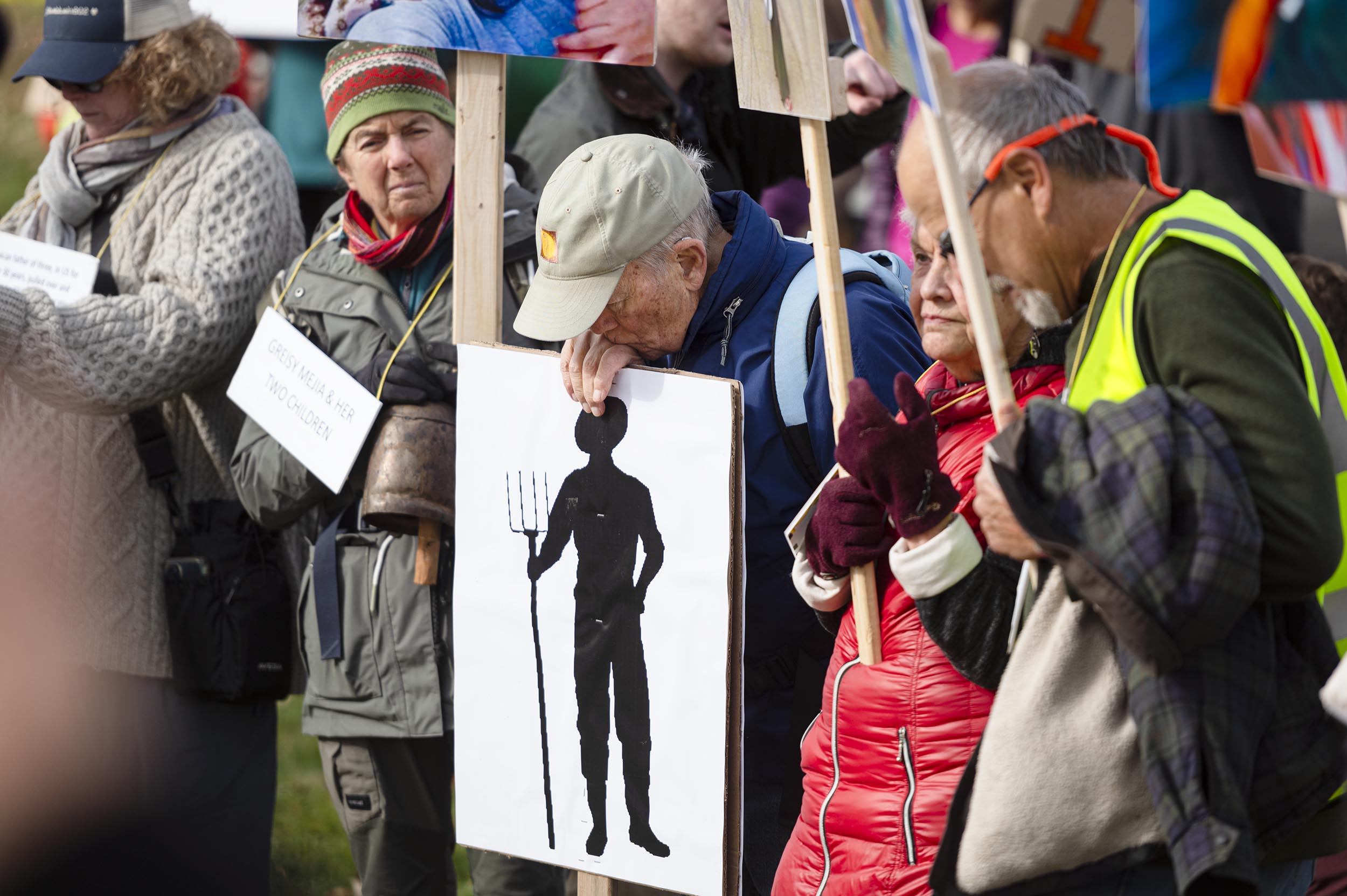 A group of people hold protest signs, including one showing a silhouette of a person with a pitchfork; they are dressed for cold weather and standing outdoors.