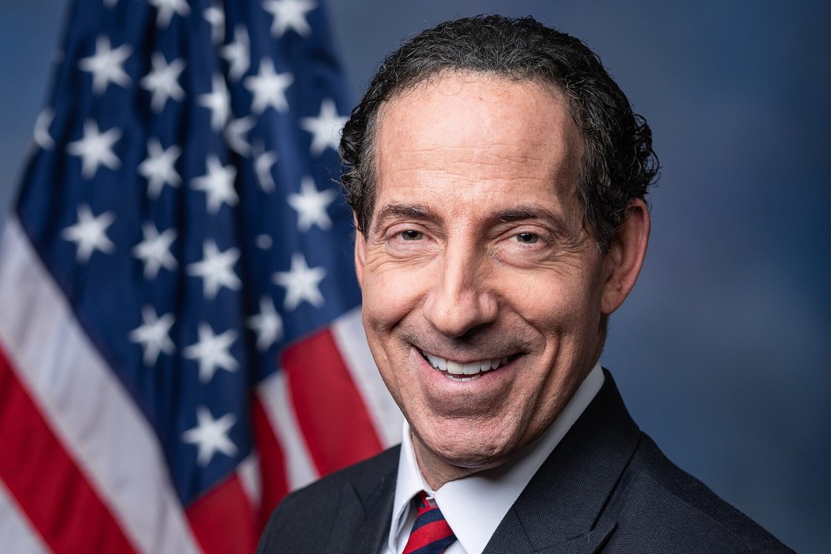 A man in a suit and tie smiles in front of a blurred United States flag backdrop.