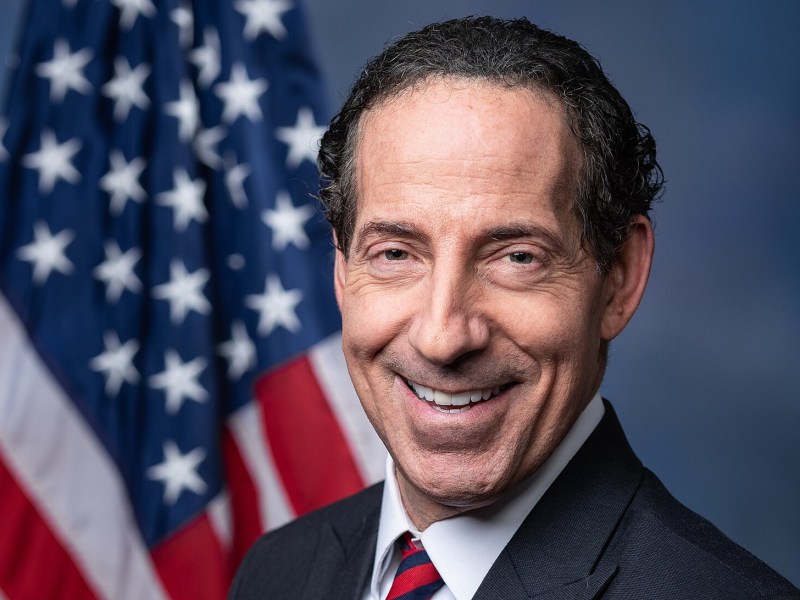 A man in a suit and tie smiles in front of a blurred United States flag backdrop.