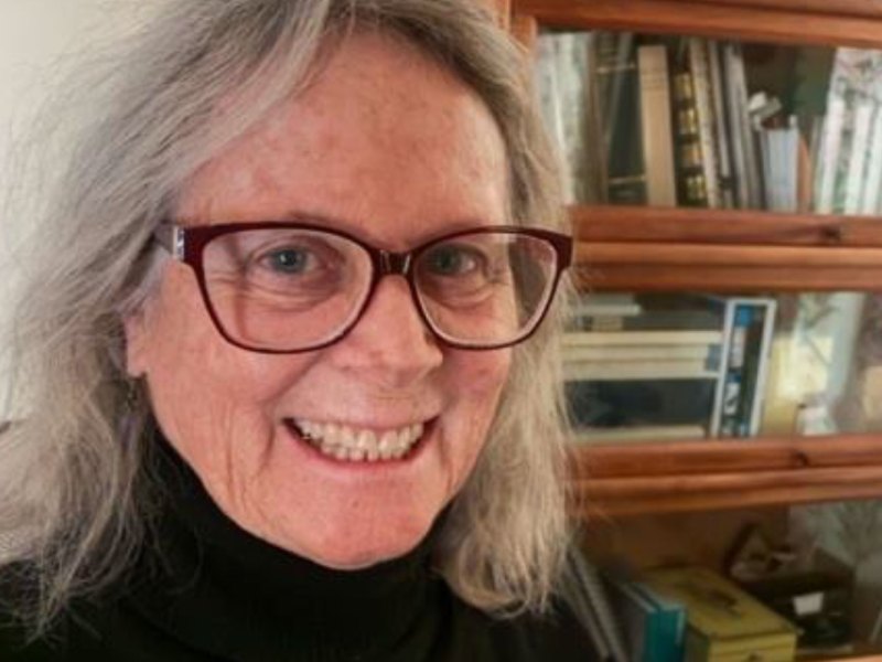 Older woman with gray hair and glasses, wearing a black turtleneck, smiling in front of a wooden bookshelf filled with books and various items.