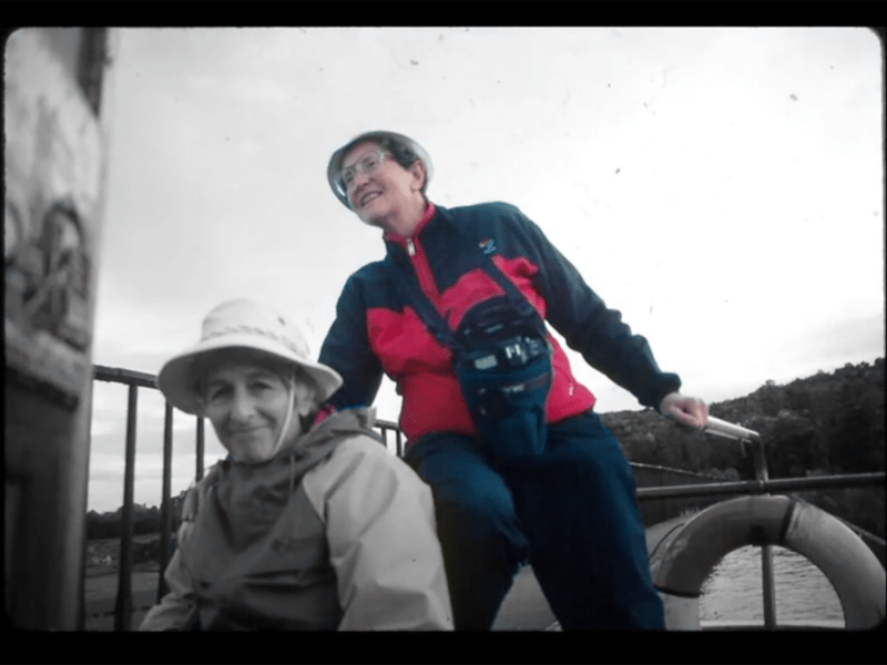 Two older adults sit and stand on a boat deck outdoors, dressed in jackets and hats, with a lake and trees visible in the background.
