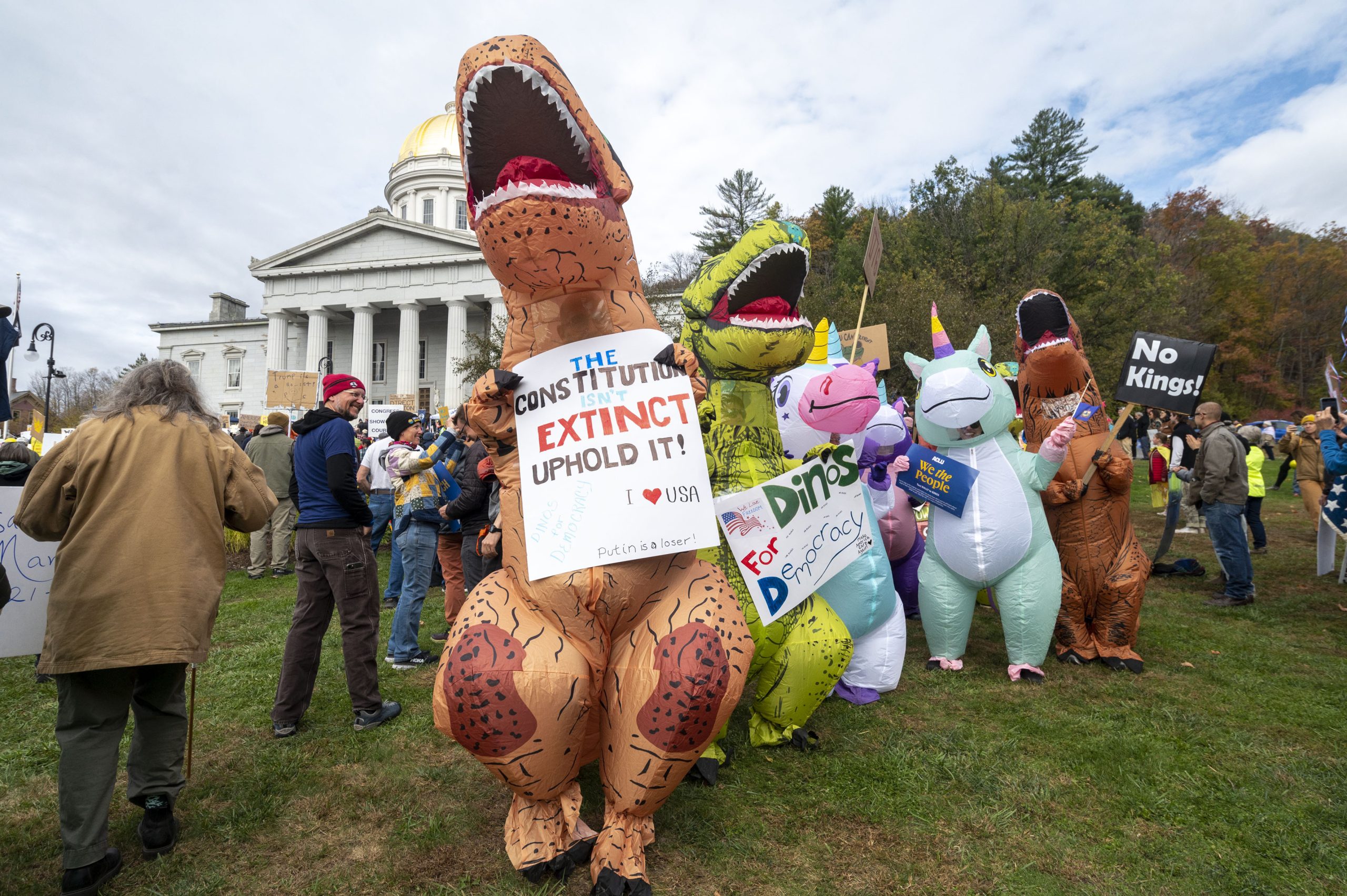 People in inflatable dinosaur costumes hold protest signs on a grassy area in front of a white-domed government building.