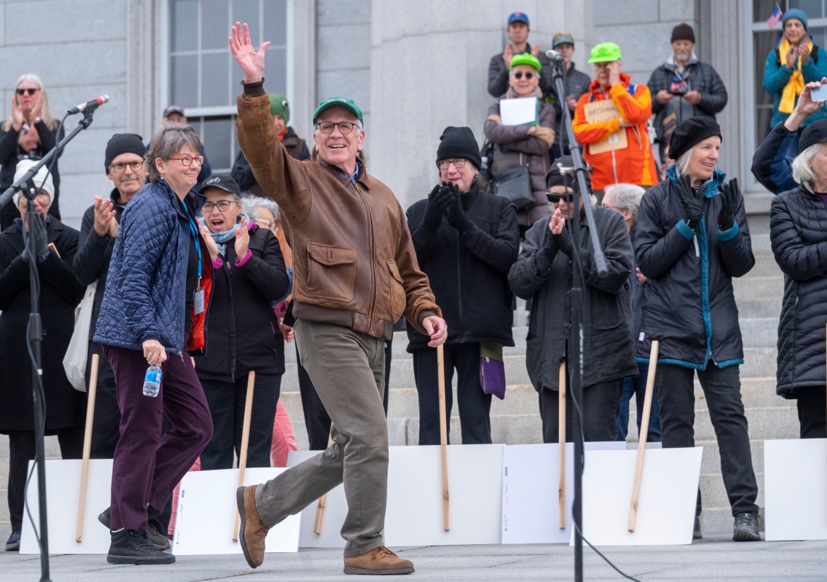A man in a brown jacket and green cap waves and smiles while walking past a group of people standing and clapping on stone steps.