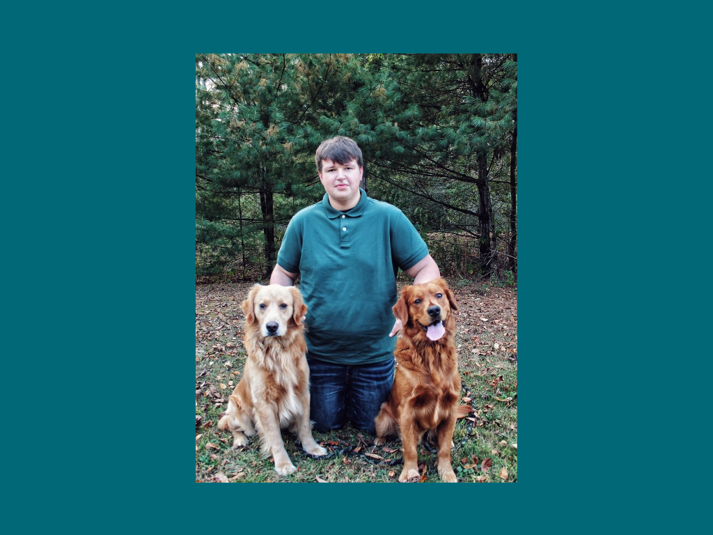 A young man kneels outdoors on grass with two golden retrievers, one on each side, in front of green trees.