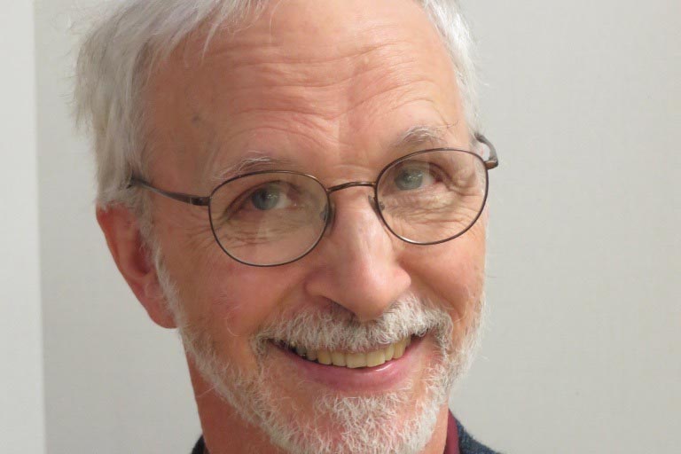 An man with a beard and glasses is smiling at the camera against a plain light background.