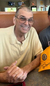 Man wearing glasses and a pale yellow polo shirt sits at a restaurant booth, smiling at the camera. A Boston Bruins hat with signatures rests on the table in front of him.