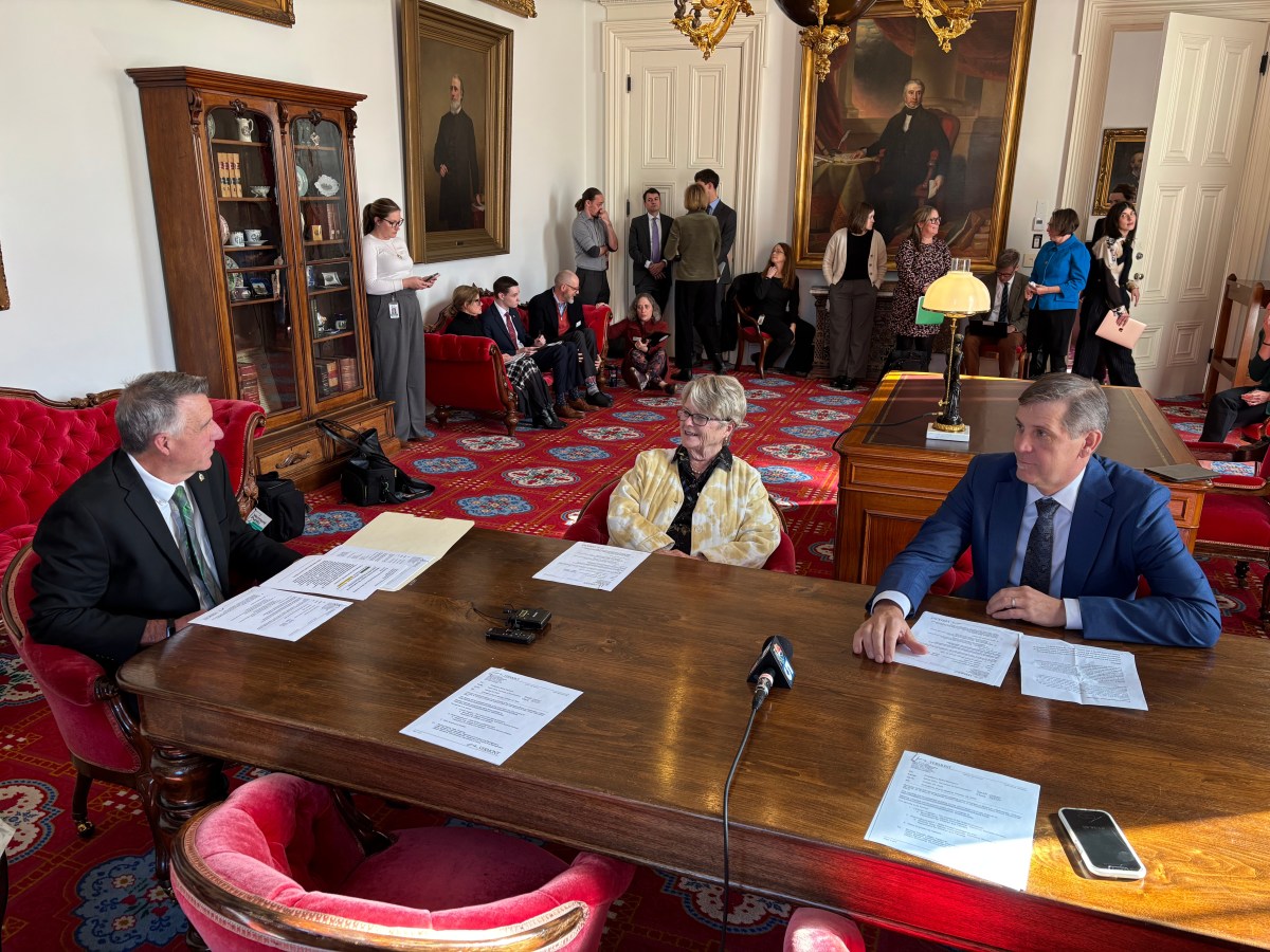 Three people sit at a large wooden table with documents in a formal room, while others observe and portraits hang on the walls in the background.