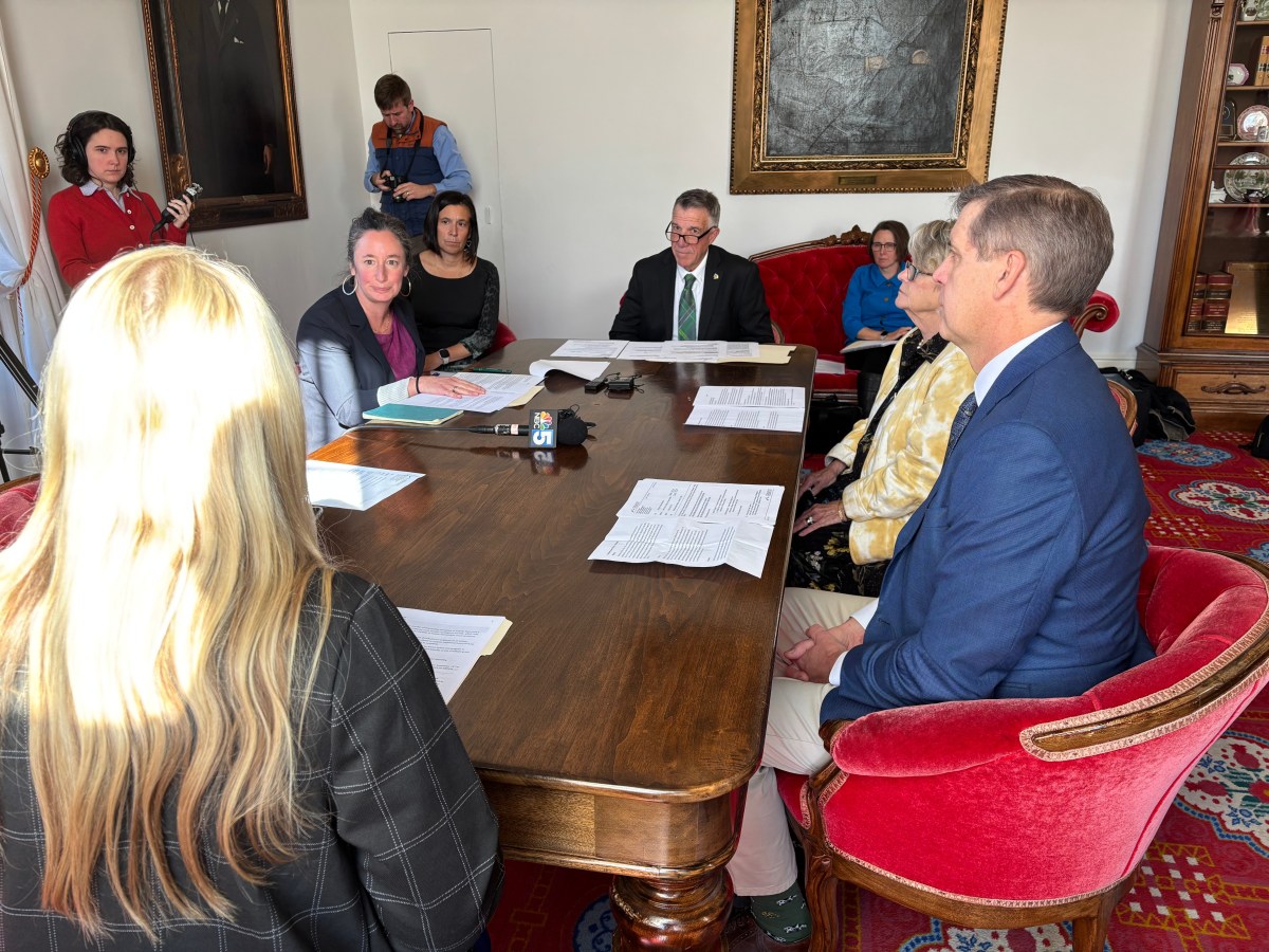 Several people sit around a wooden table in a formal room with papers in front of them, while others observe or take notes nearby.