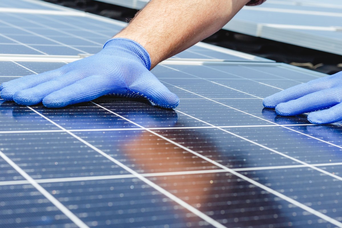 A person wearing blue gloves installs or inspects solar panels, with hands positioned on the surface of the panels.
