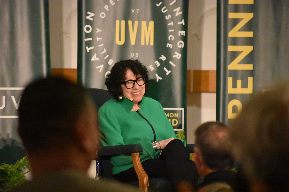 A woman in a green jacket sits and smiles onstage during a public event, with UVM banners displaying words like "innovation" and "integrity" in the background.