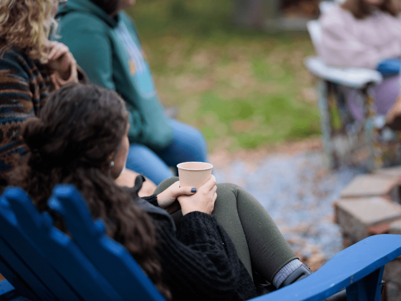 A person sits in a blue chair holding a paper cup, with others seated nearby outdoors on a grassy, leaf-strewn area.