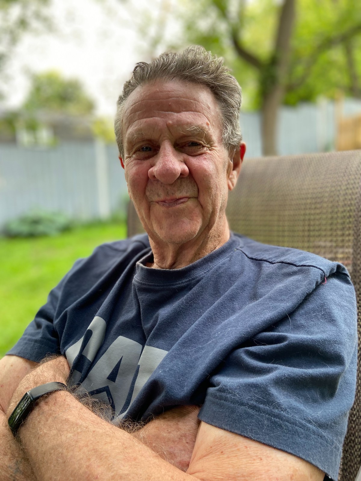 A man sitting outdoors on a cushioned chair, arms crossed, wearing a blue t-shirt and smartwatch, with a blurred yard and trees in the background.