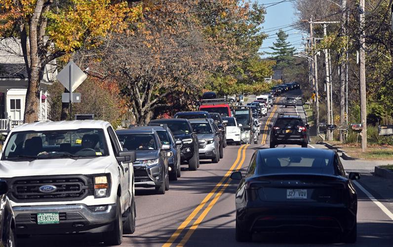 A line of cars travels in both directions on a two-lane road bordered by trees and utility poles on a clear day.