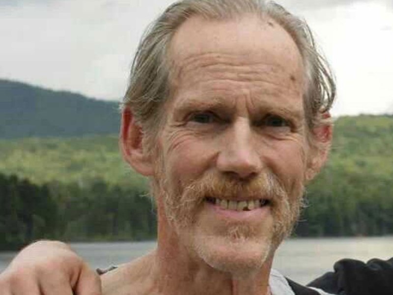 A man with light hair and a mustache stands outdoors, smiling, with a forest and lake in the background.