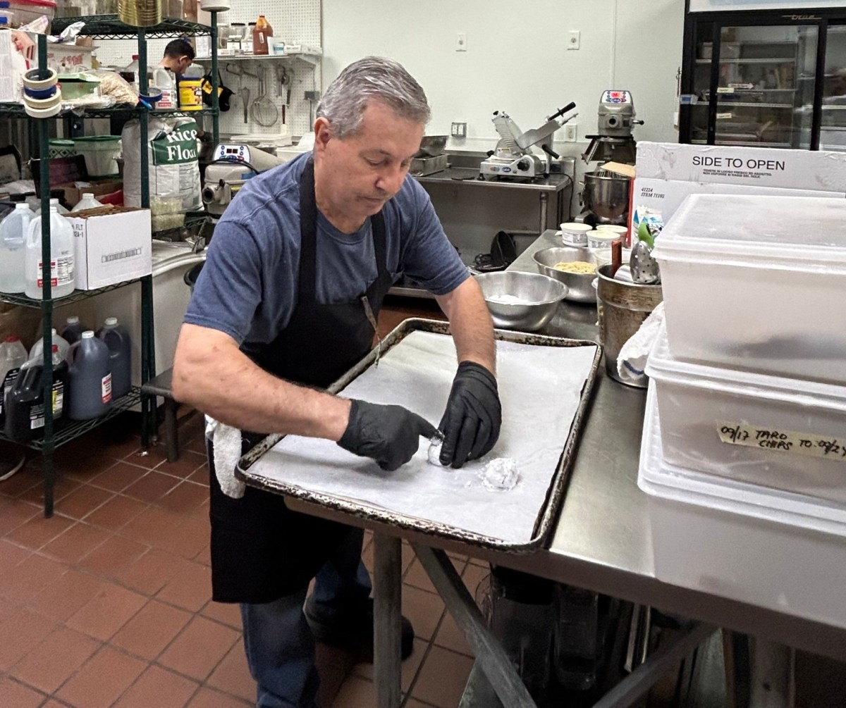 A man wearing gloves prepares food on a tray in a commercial kitchen, surrounded by ingredients and kitchen equipment.