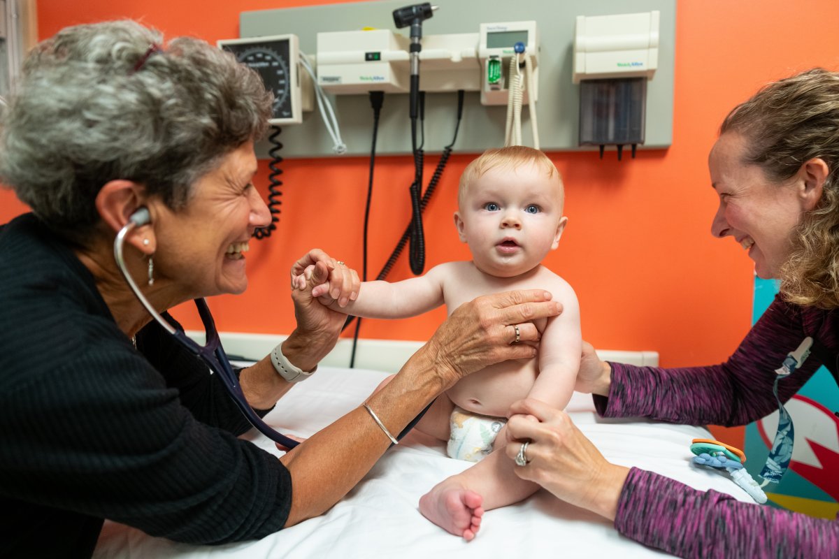 A doctor examines a baby with a stethoscope while the baby sits on an exam table and an adult supports the baby's arm in a medical office.
