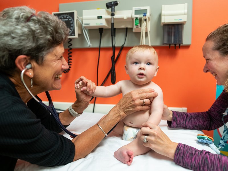 A doctor examines a baby with a stethoscope while the baby sits on an exam table and an adult supports the baby's arm in a medical office.