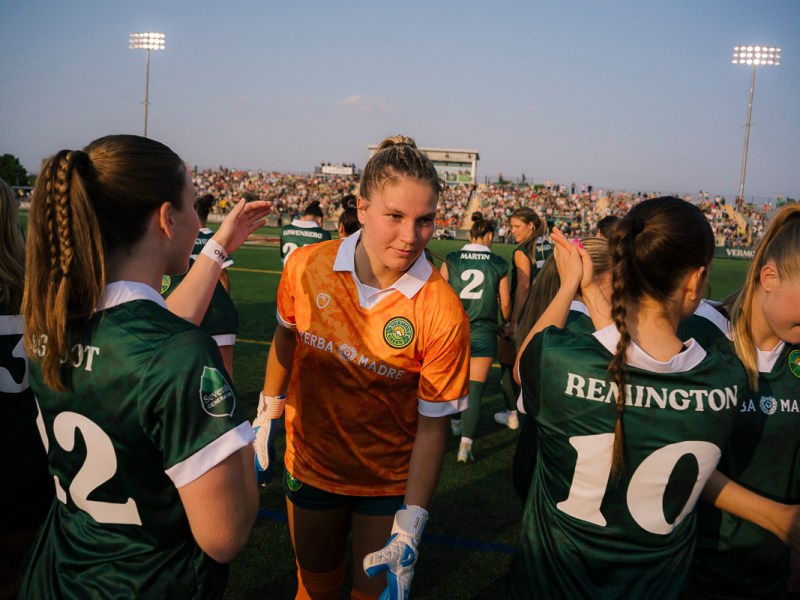 A women's soccer team in green uniforms and a goalkeeper in orange greet each other on the field, with a crowd in the background under stadium lights.