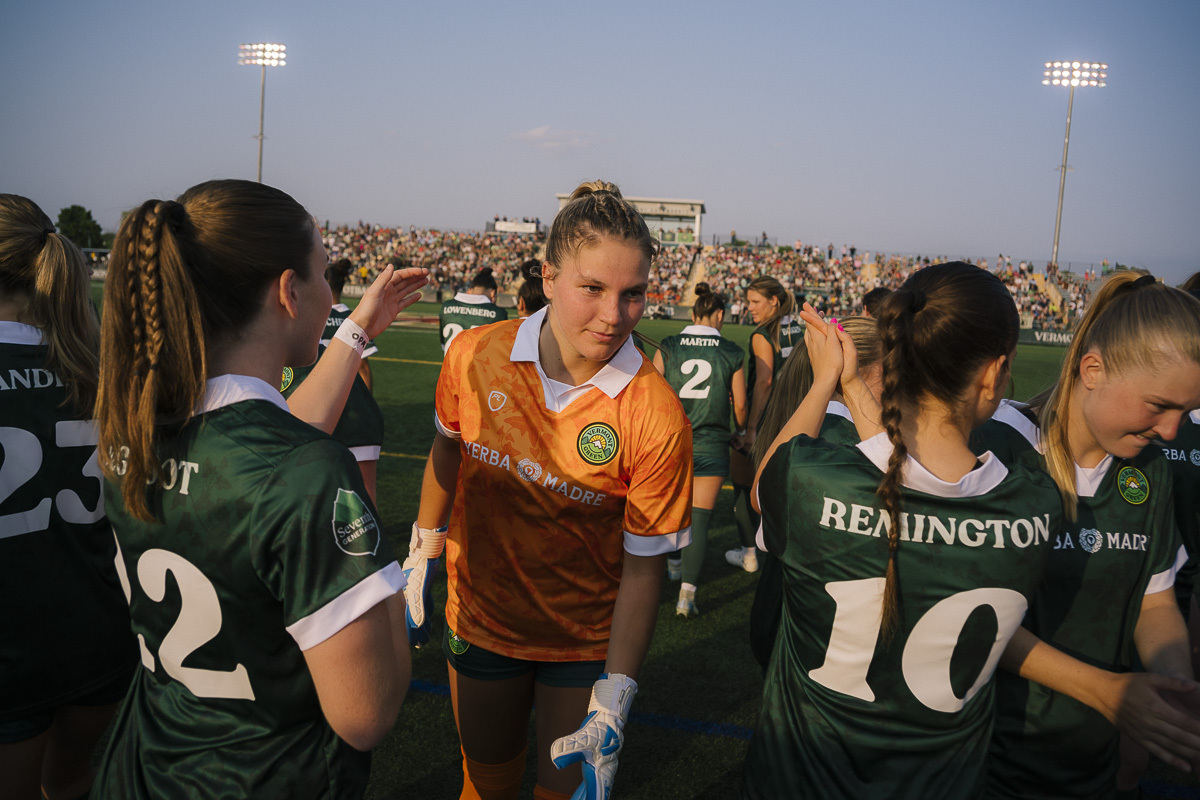 A women's soccer team in green uniforms and a goalkeeper in orange greet each other on the field, with a crowd in the background under stadium lights.