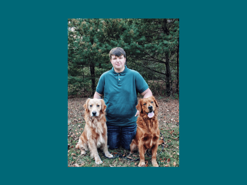 A person in a green polo shirt kneels on grass between two golden retrievers, with trees in the background.