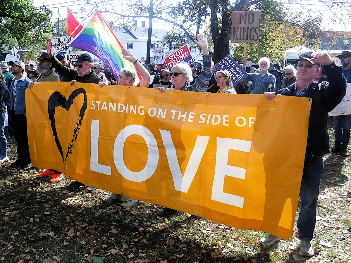 A group of people at a protest hold a large yellow banner reading "Standing on the Side of Love"; other signs and a rainbow flag are visible in the background.